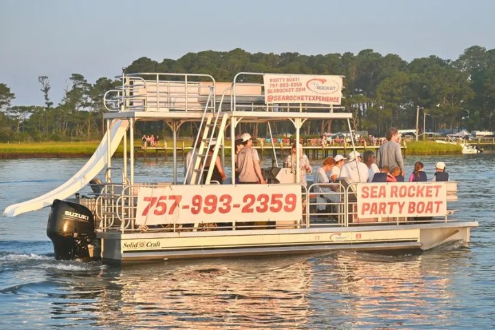 Party boat with slide on water, several people aboard, trees in background.