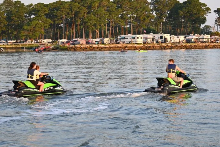 Two people on jet skis riding across a lake near a campground.