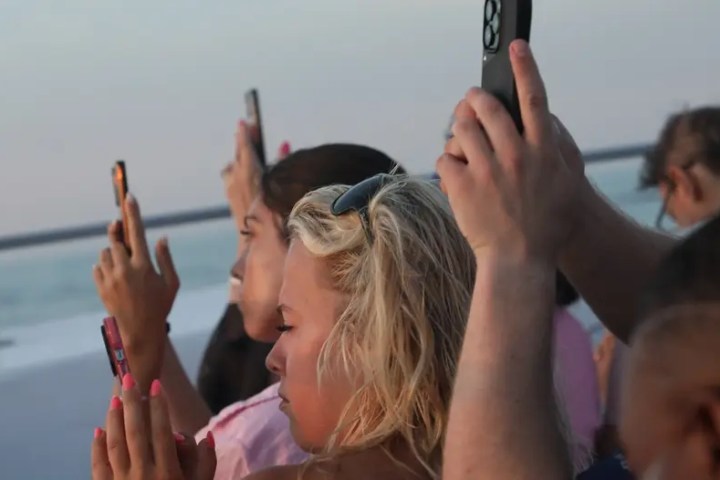 People on deck of a boat taking photos with smartphones at sunset.