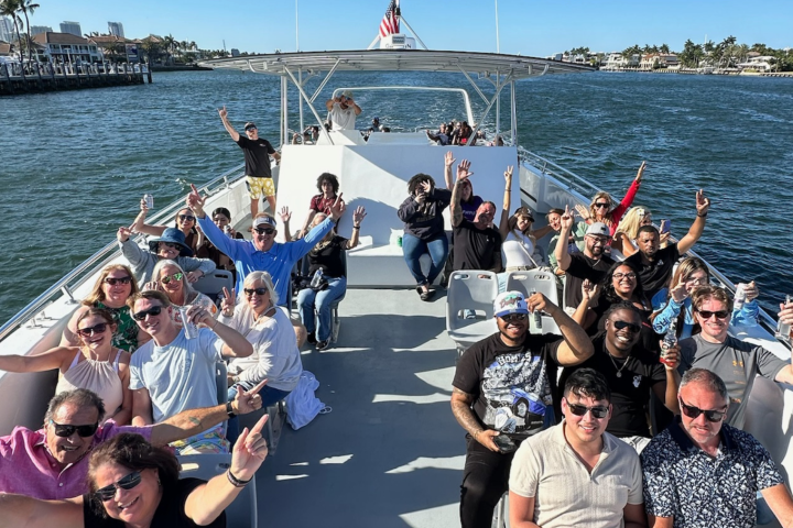 Group of people smiling and waving on a boat in sunny weather.