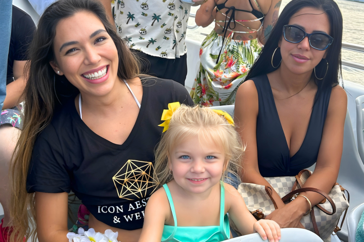 Three people smiling on a boat, including a child with a yellow bow, a woman with a black shirt, and another with sunglasses.