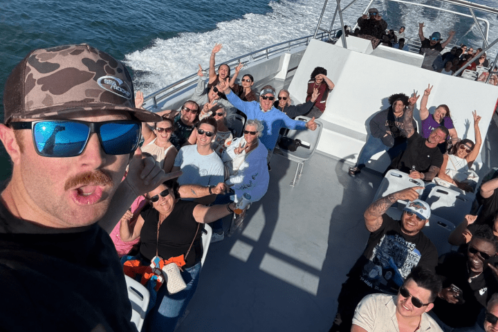 Group of people taking a selfie on a boat, smiling and waving, under clear skies.