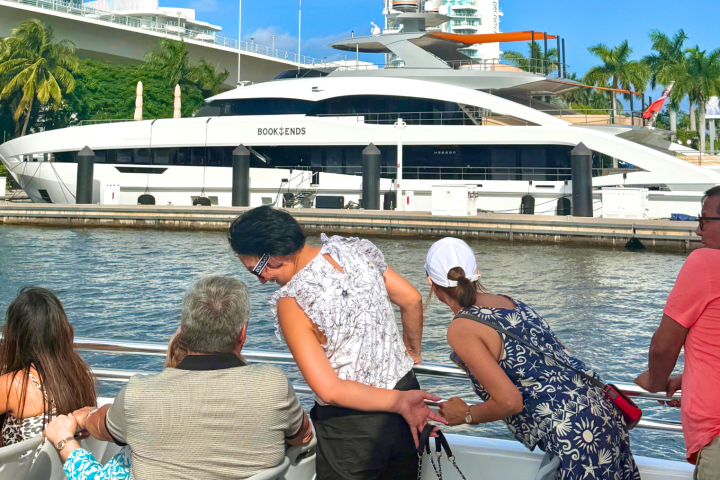 People on a boat tour observing a large yacht docked nearby under a clear blue sky.