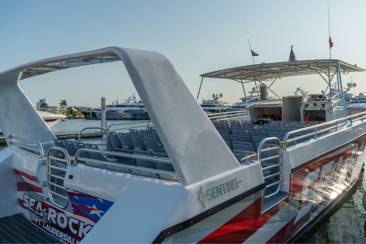 A modern, open-deck tour boat docked at a marina with empty seats and a visible cityscape in the background.