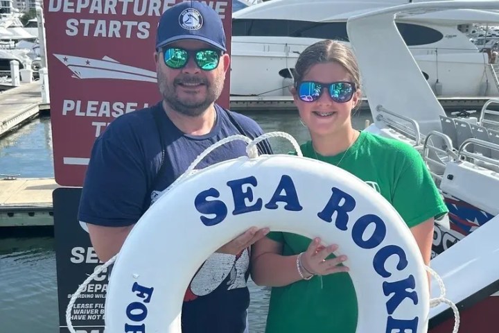 Two people holding a lifebuoy with 'Sea Rocket' near a marina with boats.