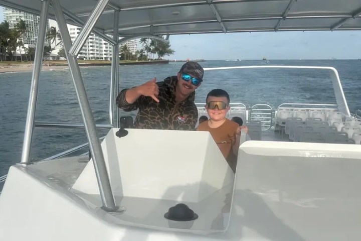 Two people smiling on a boat near a beach with buildings and palm trees in the background.