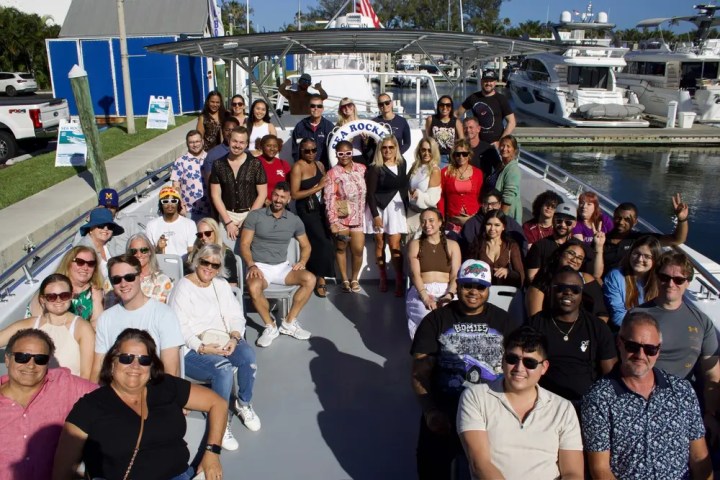 Group of people sitting on a boat, smiling and posing for a photo at a marina.