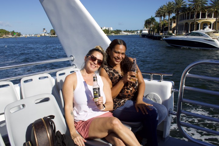 Two women smiling and holding drinks on a boat with a scenic waterfront background.
