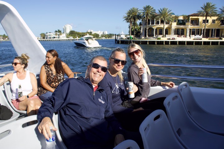 People on a boat smiling, holding drinks, with a waterfront view and palm trees in the background.