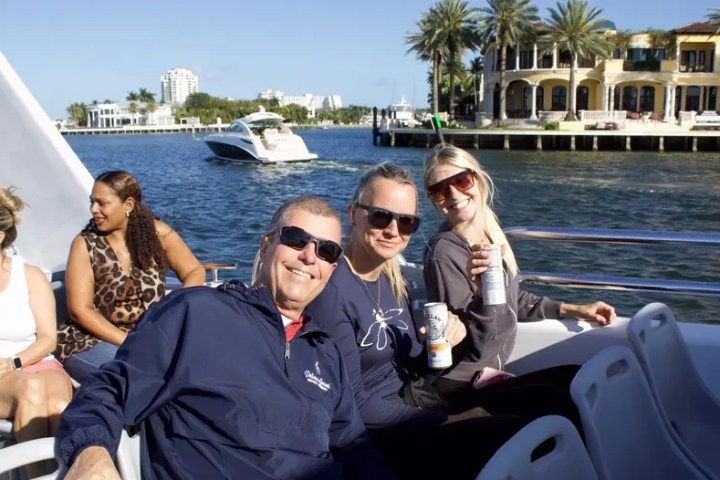 Group of people on a boat in a sunny waterfront setting with luxury homes and palm trees in the background.