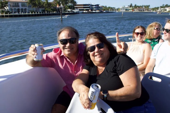 Two people smiling with drinks on a boat, sunny day, three others in the background.