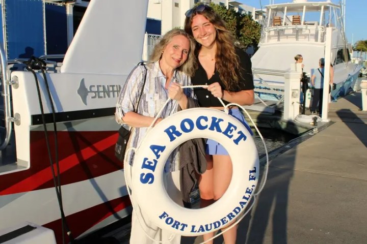 Two people smiling, holding a 'Sea Rocket' life ring at a marina.