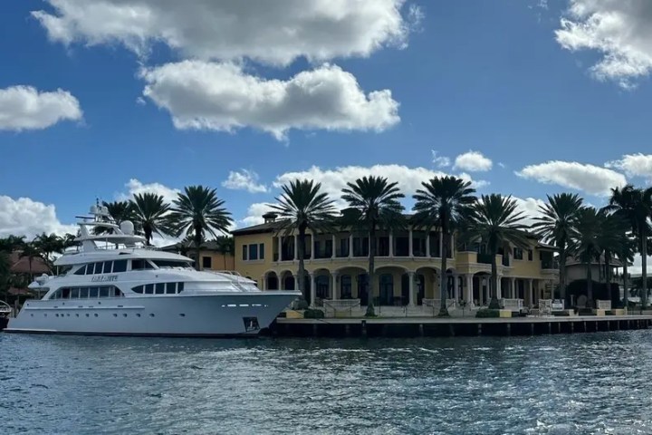 Large yacht docked by a luxurious waterfront villa with palm trees.