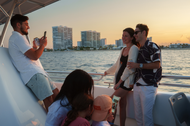 People on a boat enjoying sunset, with buildings in the background and two taking photos.