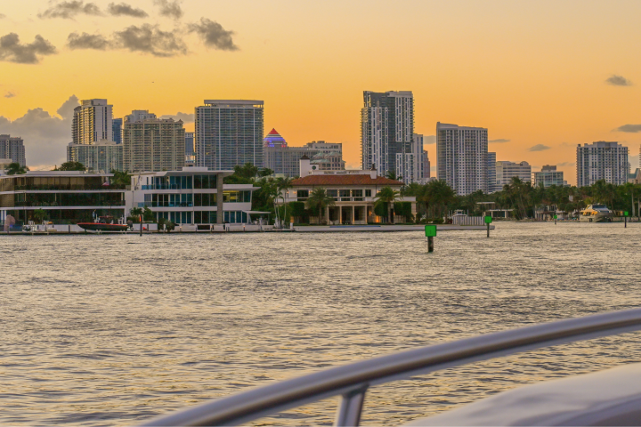 Sunset over a city skyline with modern buildings and water in the foreground.