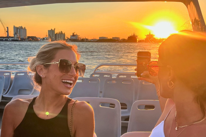 Two women on a boat at sunset, one smiling, the other taking a photo of the sun over a harbor.