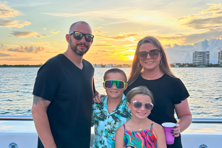 Family of four wearing sunglasses, smiling on a boat at sunset over water.