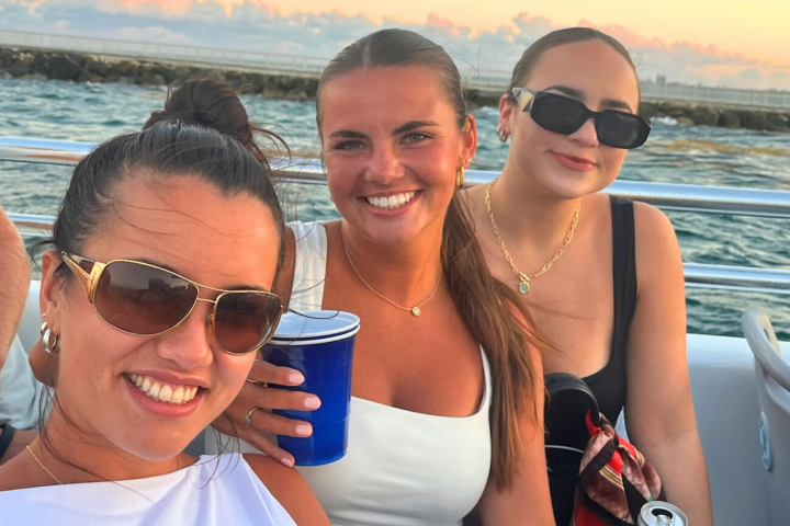 Three women smiling on a boat at sunset with ocean in background.