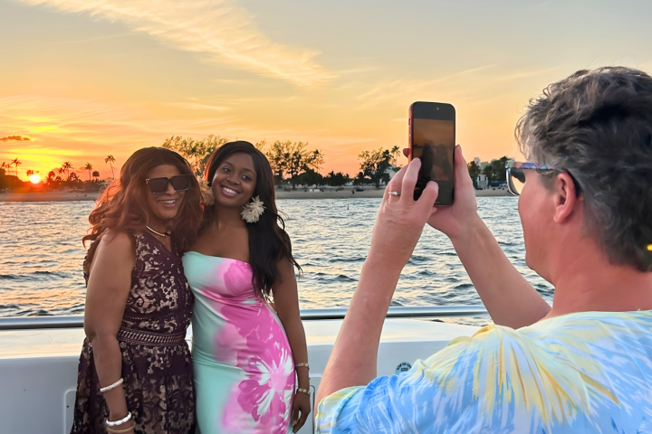 Two women posing on a boat at sunset, while a third person takes their photo with a smartphone.