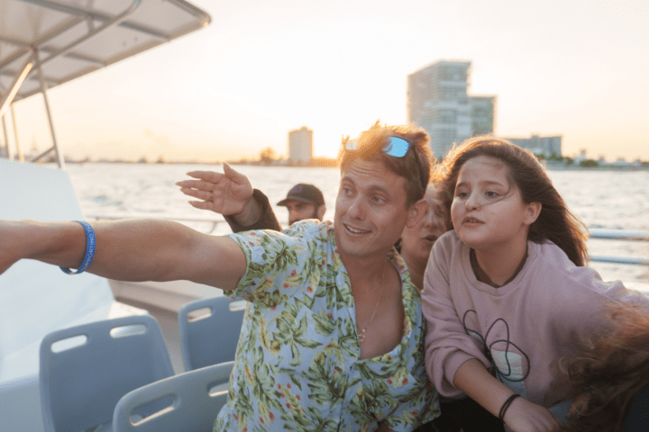 People on a boat at sunset look and point towards the water with buildings in the background.