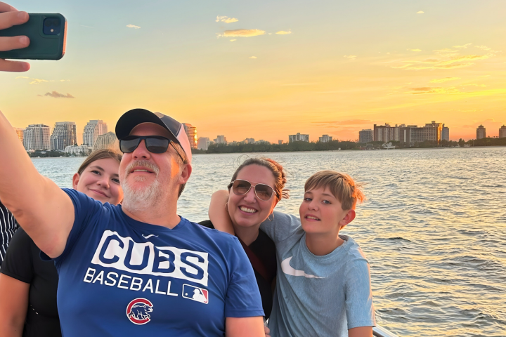 Family taking a selfie on a boat at sunset with city skyline in the background.