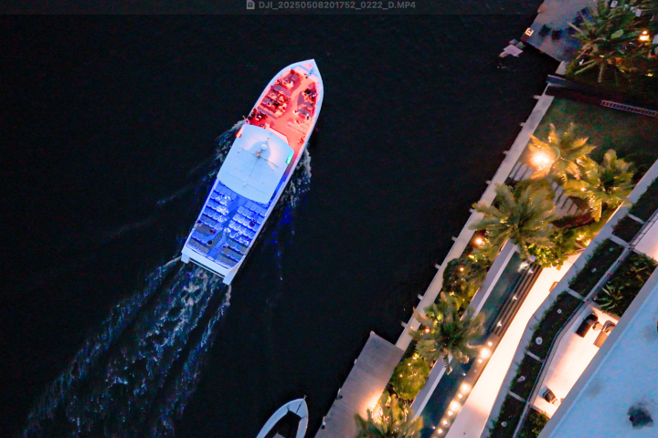 Aerial view of a boat with red and blue lights on a dark river next to a lit, palm-lined pathway.