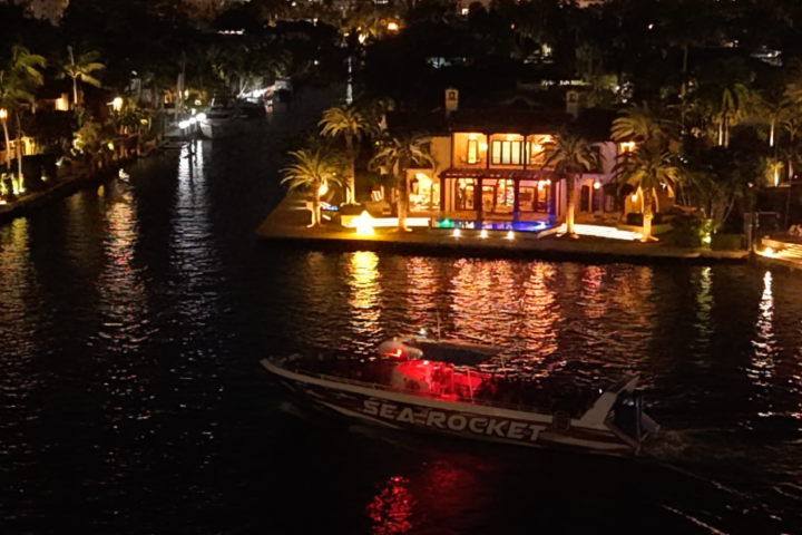 Lit boat on a waterway, passing a brightly lit house at night with palm trees around.