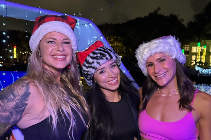 Three women in festive hats smiling at night with holiday lights in the background.