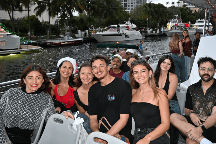 Group of people smiling on a boat with decorated boats and palm trees in the background.