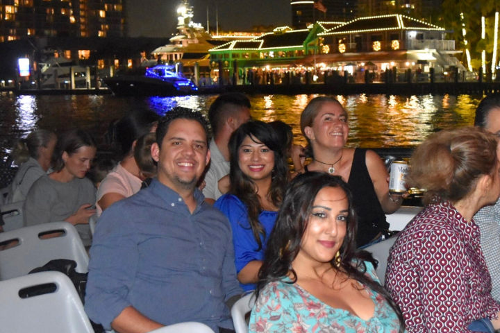 Group of people smiling on a boat at night with lit buildings in the background.