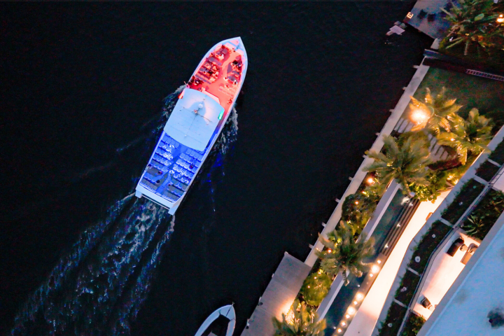 Aerial view of boat with colorful lights sailing on dark water near illuminated buildings.