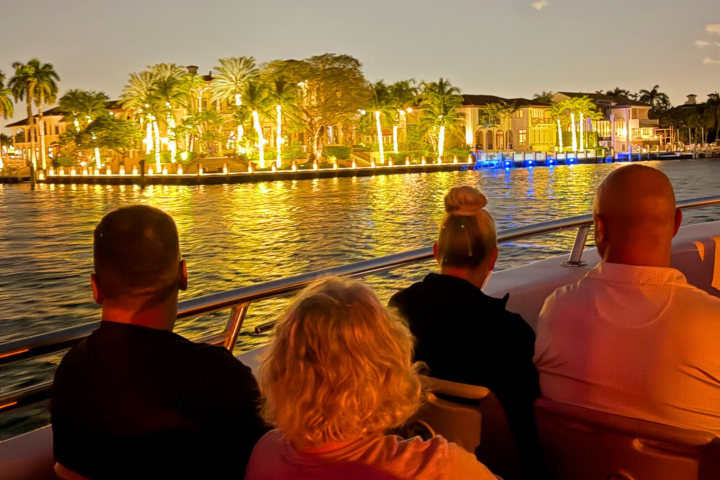 Four people on a boat at dusk, viewing illuminated waterfront homes and palm trees.
