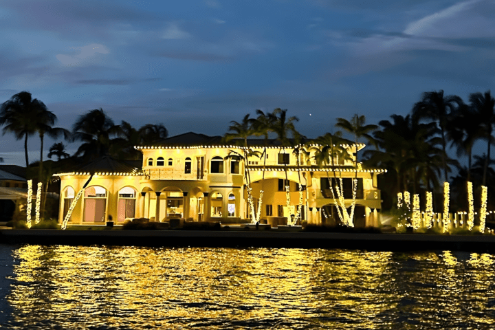 Illuminated waterfront mansion with palm trees at night.