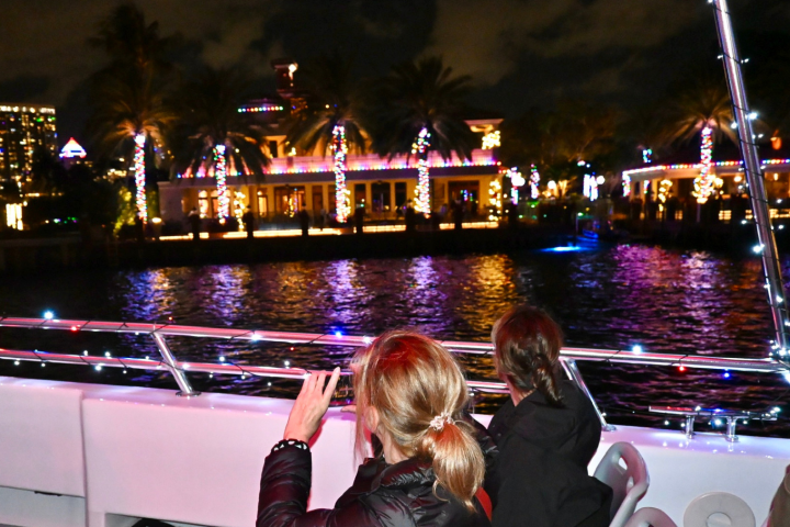 People on a boat admire a festive, illuminated waterfront with palm trees and colorful lights at night.