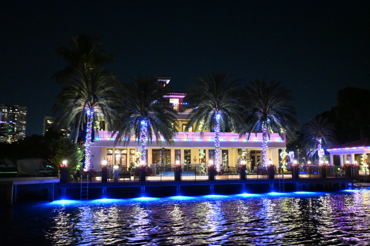 Brightly lit house with palm trees and colorful lights by the water at night.