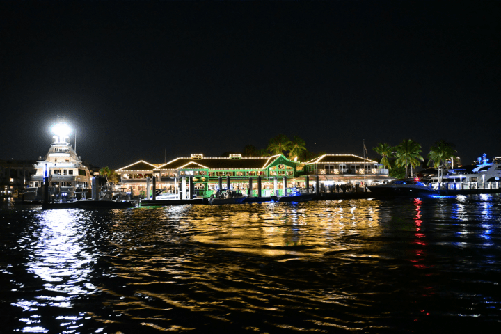 Waterfront restaurant lit at night with palm trees and yachts.
