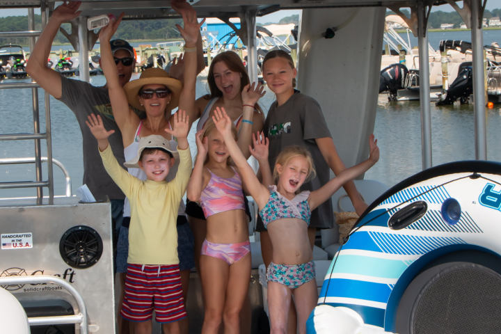 Group of adults and children smiling on a boat with arms raised, next to an inflatable tube.