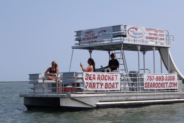 Party boat with people on a calm body of water, featuring banners and a shaded upper deck.
