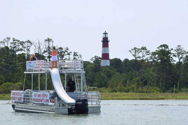 Pontoon boat with a slide on water, lighthouse in background, trees along the shore.
