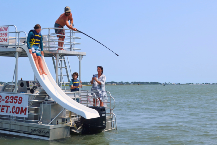 Children slide off a boat slide into the water; two adults watch, one fishing with a rod.
