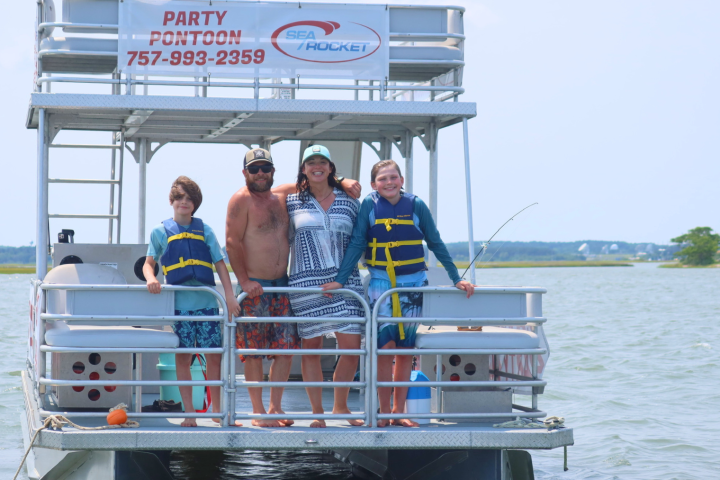 Group on pontoon boat, smiling, wearing life jackets, on calm water.