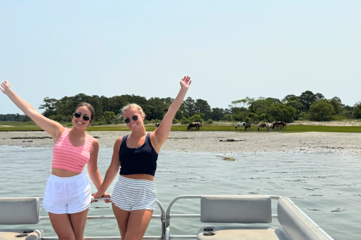 Two people on a boat waving with horses on a distant shore.
