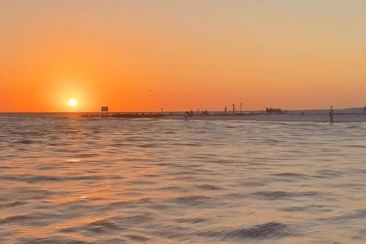 Sunset over a calm ocean with people on a distant pier and beach.