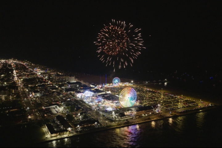 Night aerial view of amusement park with lit Ferris wheel and fireworks above.
