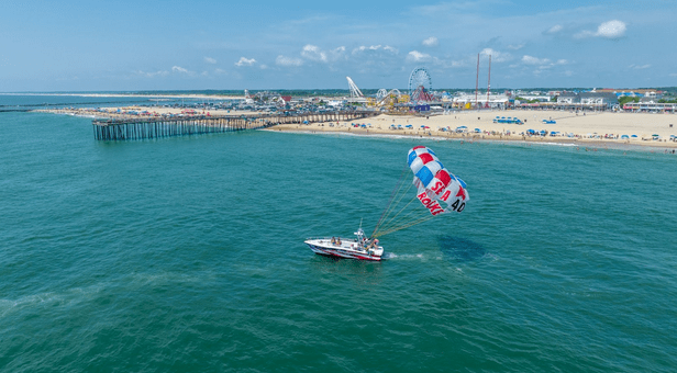 Boat parasailing near a pier with a beach, ferris wheel, and amusement park in the background.