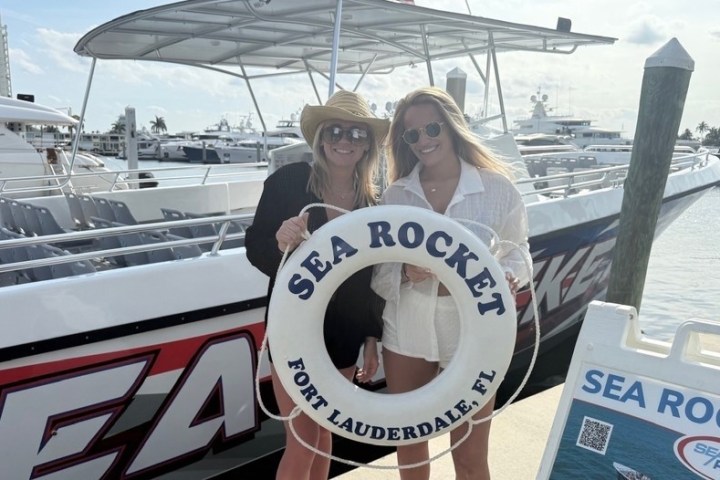 Two women holding a Sea Rocket life ring in front of a boat at a marina.