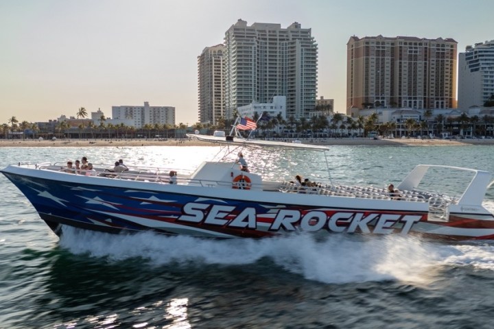 Speedboat named 'Sea Rocket' with passengers, cruising near a city shoreline with tall buildings in the background.