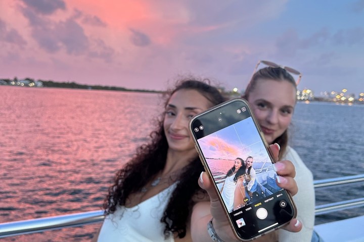Two people on a boat at sunset, one holds a phone showing their selfie against a pink sky.