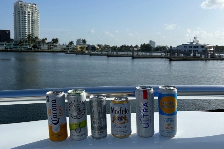 Six beverage cans on a boat railing with a waterfront cityscape in the background.