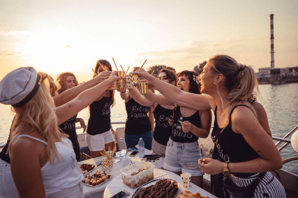Group of women toasting with drinks on a boat at sunset, wearing matching attire.